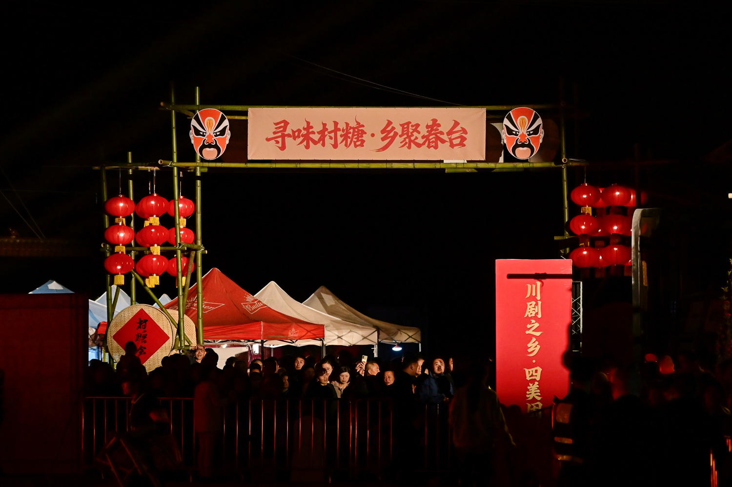 2026年固驿春台会（村糖会）夜景主入口实拍，含“寻味村糖·乡聚春台”横幅、川剧脸谱装饰、红灯笼阵列、人群与帐篷摊位，专业夜间商业活动纪实摄影案例