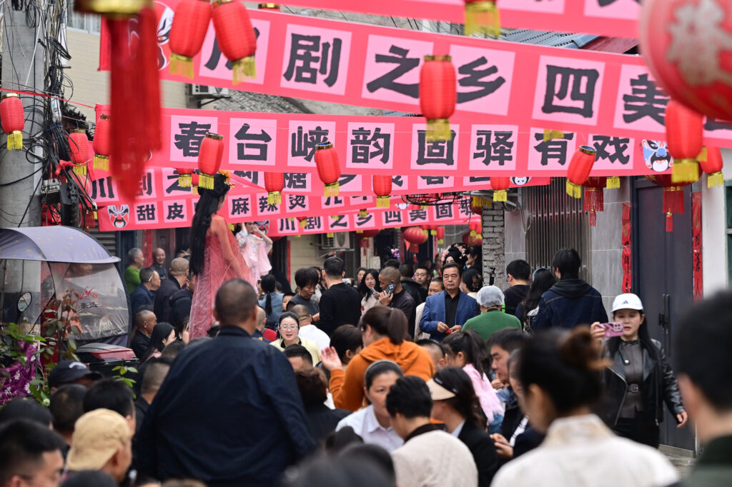 四川成都邛崃固驿街道2026年春台会非遗民俗活动现场，人潮涌动的街道悬挂“川剧之乡”横幅，摄影师记录“村糖会”双IP联动的热闹集市与传统美食摊位，人文纪实摄影