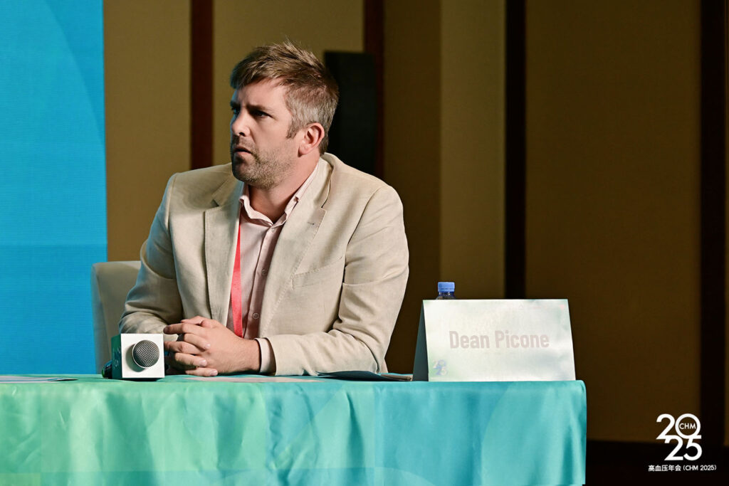 2025高血压年会(CHM 2025)特邀演讲嘉宾Dean Picone在论坛 Panel 环节发言瞬间，身着浅色西装搭配粉色衬衫，面前摆放麦克风与姓名牌，背景为蓝色LED屏幕，专业会议纪实摄影案例