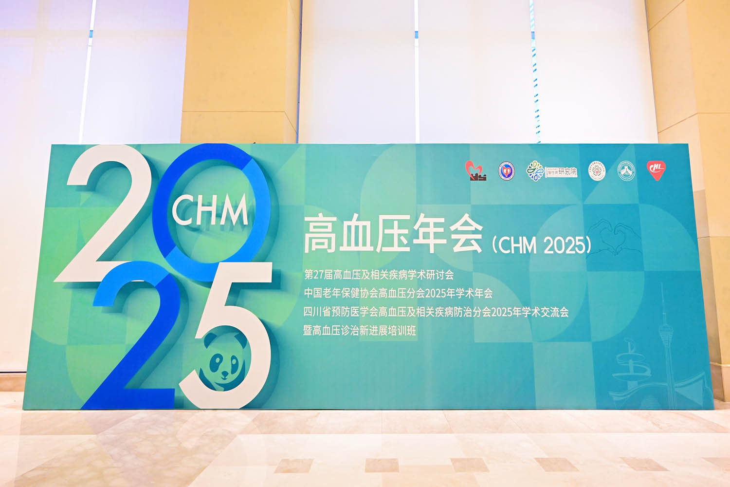 2025高血压年会(CHM 2025)主视觉背景板实拍，含“2025”立体字样、熊猫吉祥物、中国老年保健协会/四川省预防医学会等主办机构Logo，蓝绿渐变几何图案设计，专业会展摄影案例