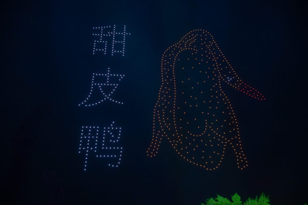 甜皮鸭主题无人机灯光秀夜景实拍，左侧白色光点组成“甜皮鸭”文字，右侧橙红色光点勾勒鸭子轮廓，专业商业活动科技视觉营销摄影案例
