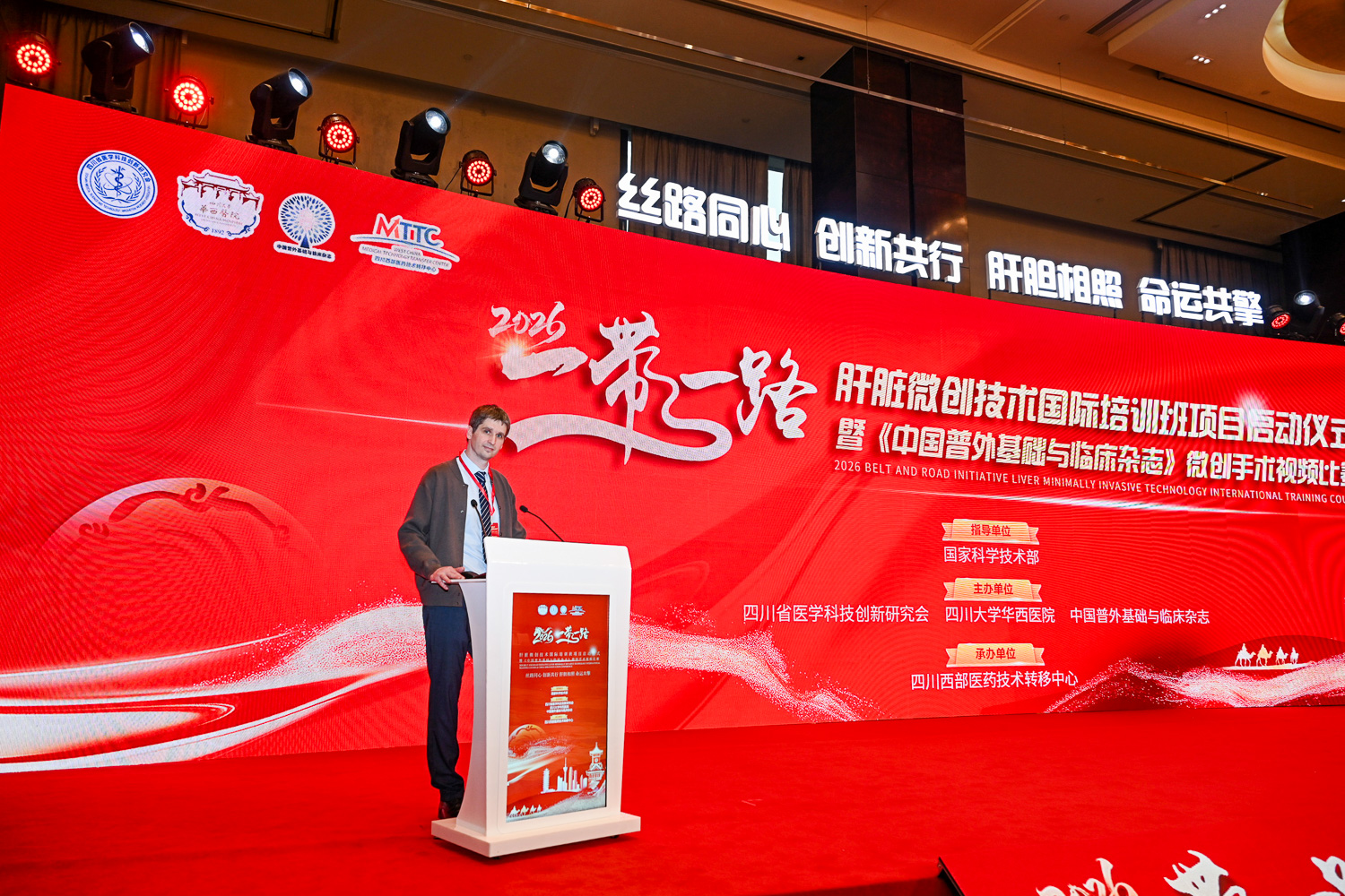 2026年一带一路肝脏微创技术国际培训班启动仪式现场，外籍专家在讲台发表演讲，身后红色背景板展示丝路同心创新共行主题，四川大学华西医院主办，成都高端医疗学术会议演讲抓拍摄影案例
